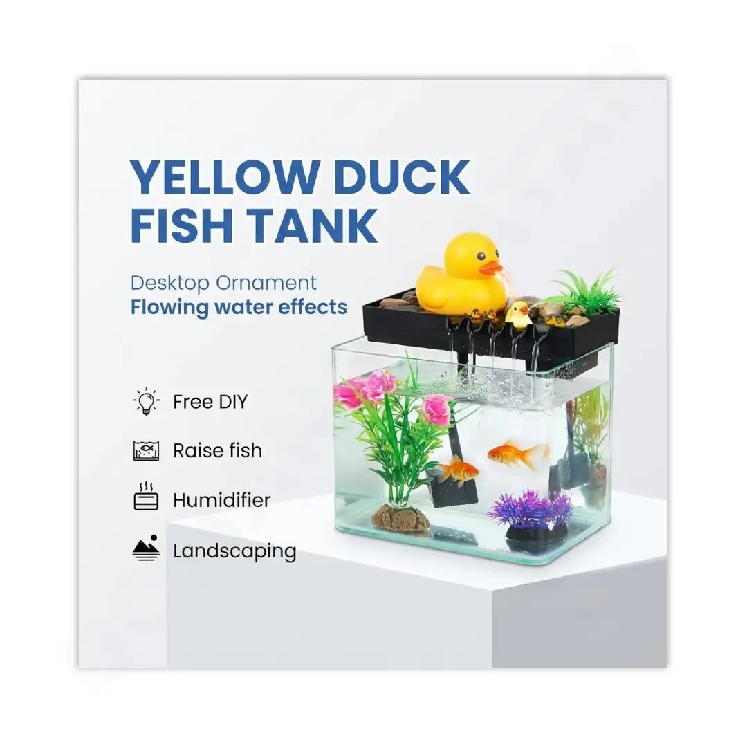 A yellow duck fish tank desktop ornament shown on a white background with promotional text. The clear rectangular tank contains water, artificial plants, rocks, and a small orange fish, with a black lid and a yellow rubber duck on top.